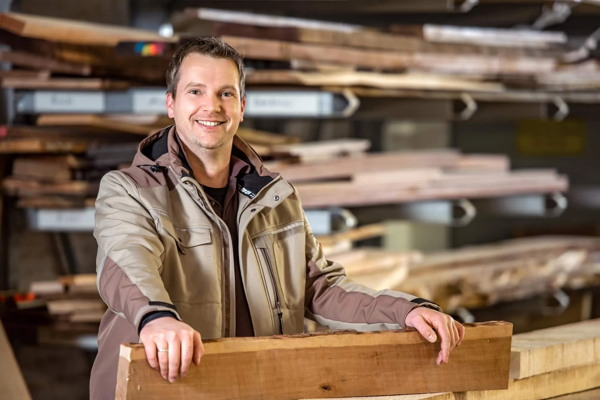 Lächelnder Schreiner in Holzlager mit Brettern und Holzbalken im Hintergrund