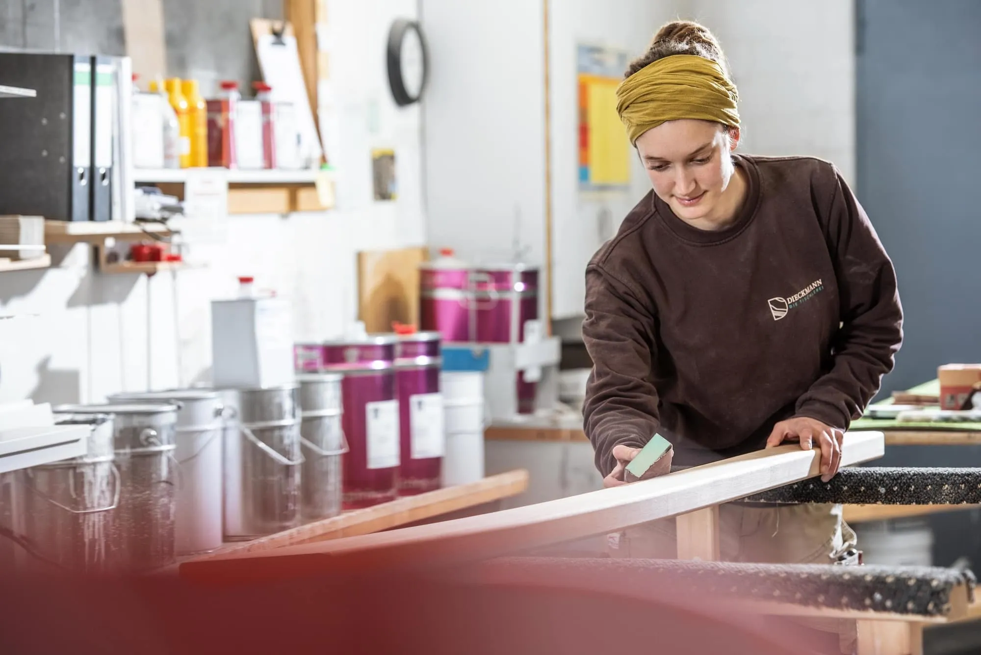 Malerin schleift Holzleiste in Werkstatt, Lackeimer und Werkzeug im Hintergrund